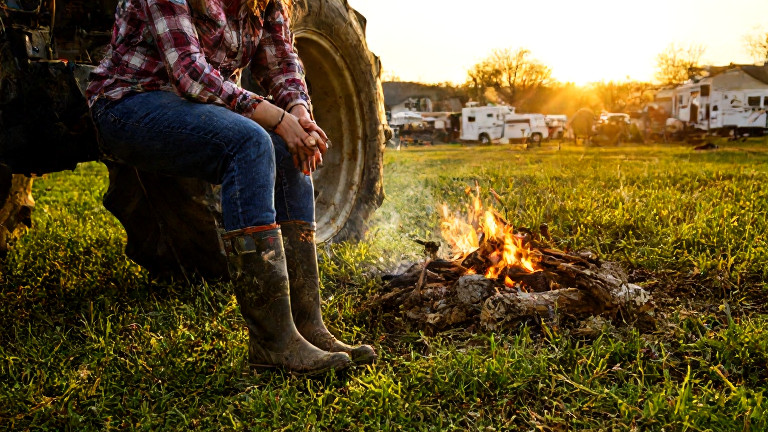 woman farmer in muddy boots leaning on tractor at sunrise