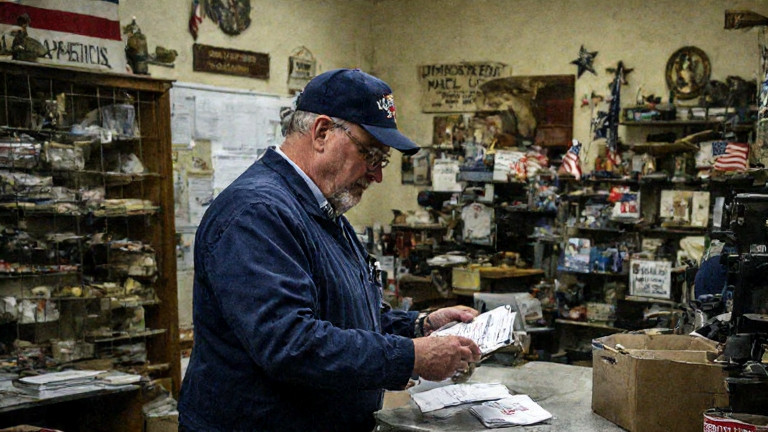 postal worker in vintage uniform carefully sorting mail in a small Vermilion post office with a "Patriot Mail" stamp on the counter