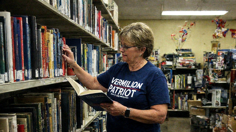 librarian in a "Vermilion Patriot" t-shirt placing a book titled "Patriot's Guide to Baghdad" on a shelf