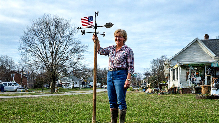 woman farmer in muddy boots holding a wooden weather vane with a small American flag attached, smiling at a partly cloudy sky