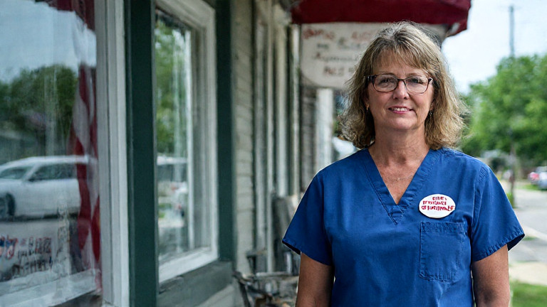 nurse in scrubs with "FREE SPEECH SARDINE" pin standing near Vermilion City Council building