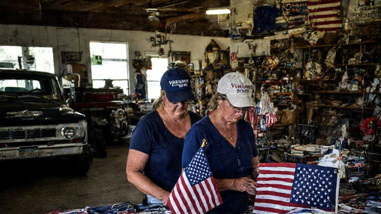 woman wearing a "Vermilion Patriot" cap examining a vintage American flag in a cluttered local shop