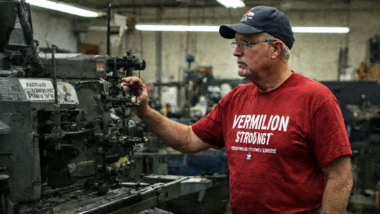factory worker in a 'Vermilion Strong' t-shirt inspecting a machine labeled 'AMERICA FIRST' at a local manufacturing plant