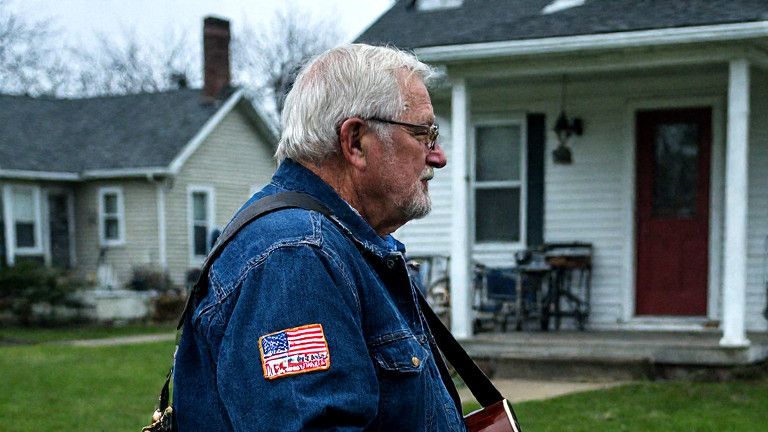 elderly veteran in a faded Marine jacket with a "Lake Erie Freedom Chorus" patch on his backpack standing by a vintage guitar case