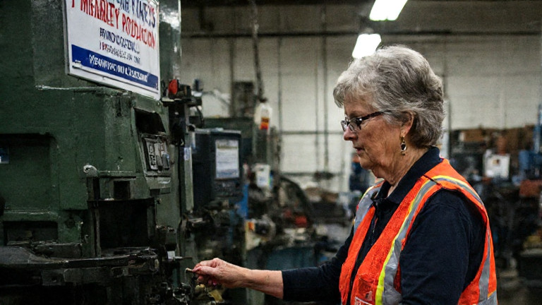 middle-aged factory worker in safety vest adjusting a machine at Vermilion Steel Plant with "Patriot Production" written on a nearby sign