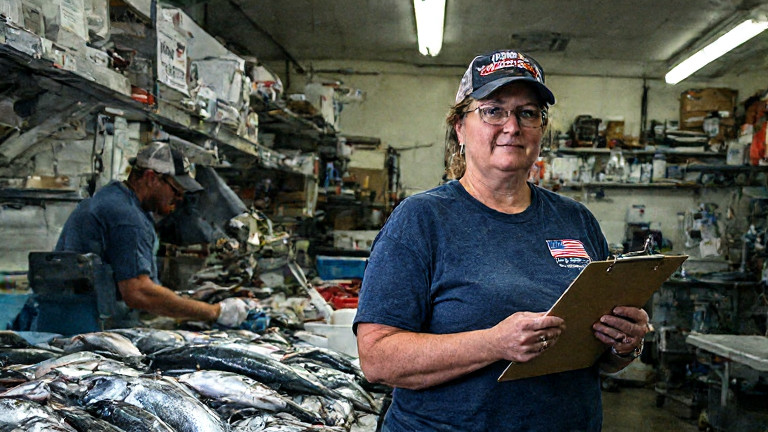 factory worker in a Vermilion Seafood Processing Co. t-shirt counting fish with a flag-emblazoned clipboard