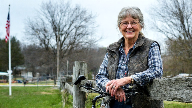 woman farmer in mud-streaked boots leaning on a tractor, smiling at a bicycle chain hanging from a fence